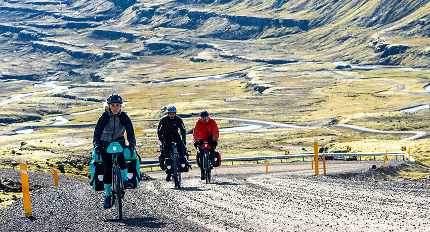Trois voyageurs à vélo équipés de sacoches latérales Ortlieb en plein ascension sur une piste de terre dans un paysage montagneux grandiose.