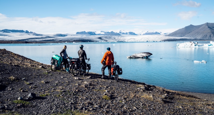 Trois cyclistes équipés de sacoches latérales contemplent un lac dominé par un glacier.