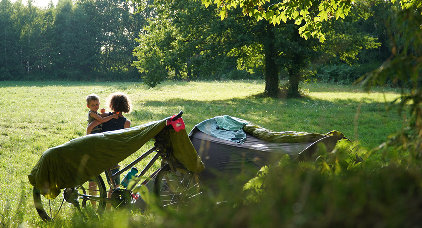 Une femme avec son petit garçon dans les bras achève de monter un bivouac en pleine nature