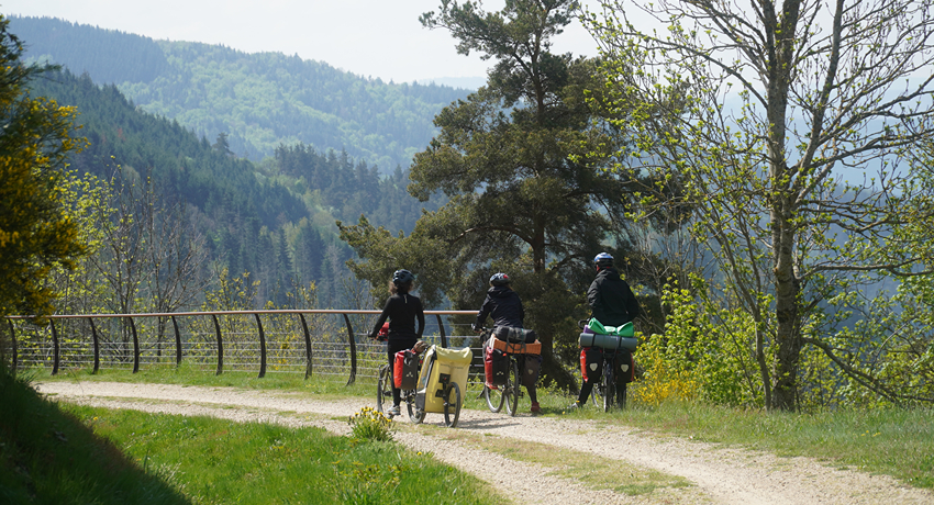 Trois randonneurs à vélo arrêtés sur une piste de terre contemplent un paysage de moyenne montagne.