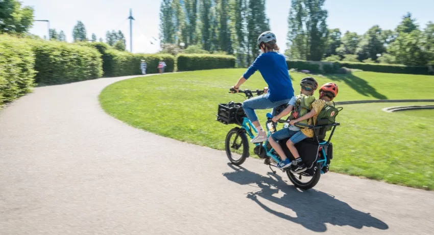 Une mère traverse un parc avec ses deux fils assis sur la plateforme d'un vélo rallongé Tern GSD.