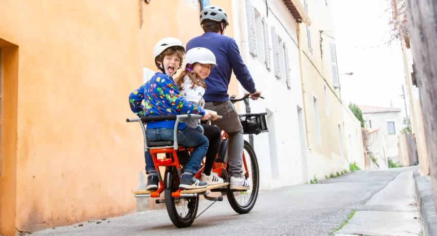 Deux enfants derrière leur père sur la plateforme arrière d'un vélo rallongé Yuba Spicy Curry.