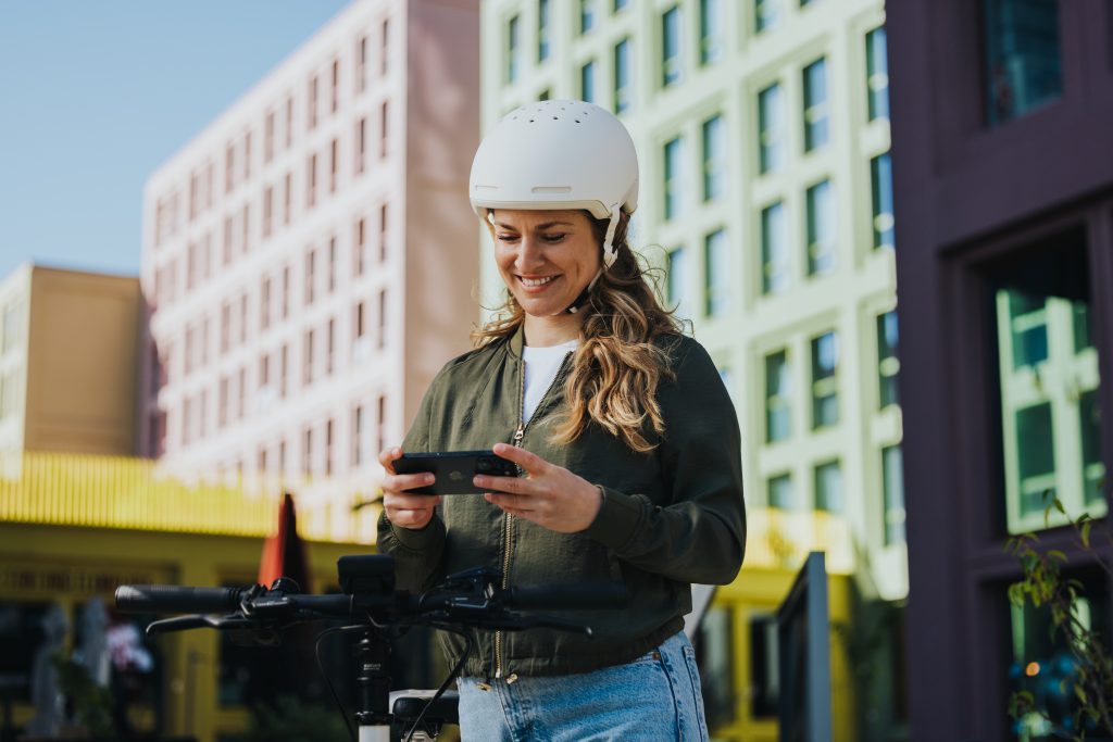 Devant, une rangée d'immeubles, une cycliste regarde son smartphone dans lequel est consigné le carnet d'entretien de son vélo électrique.