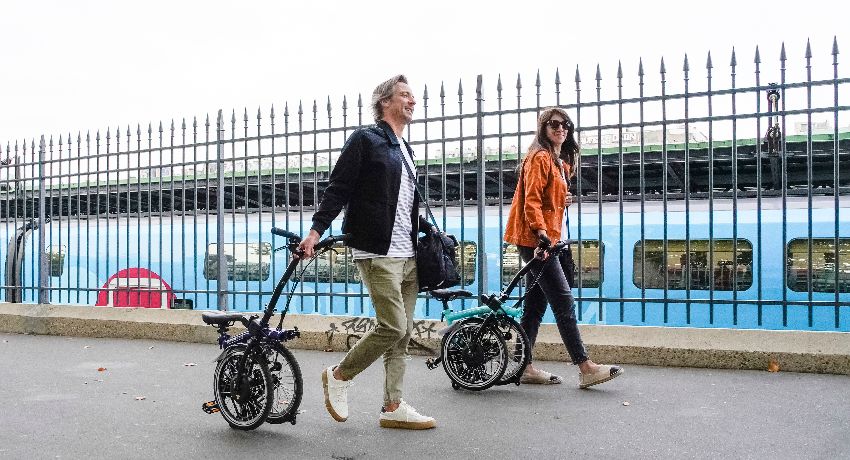 Un homme et une femme marchent sur un quai de gare en tirant leur vélo pliant comme une valise à roulettes.