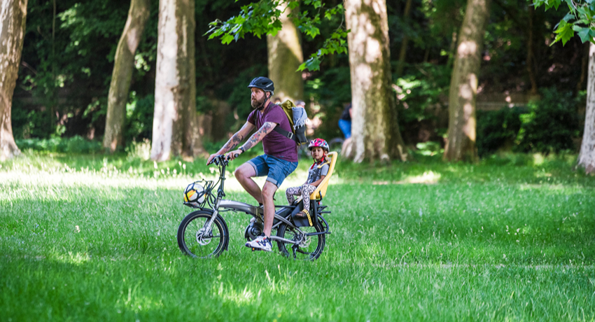Un père transporte son enfant sur un siège enfant installé à l'arrière d'un vélo pliant.