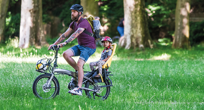 Un père transporte son fils sur un siège enfant installé à l'arrière d'un vélo pliant. 