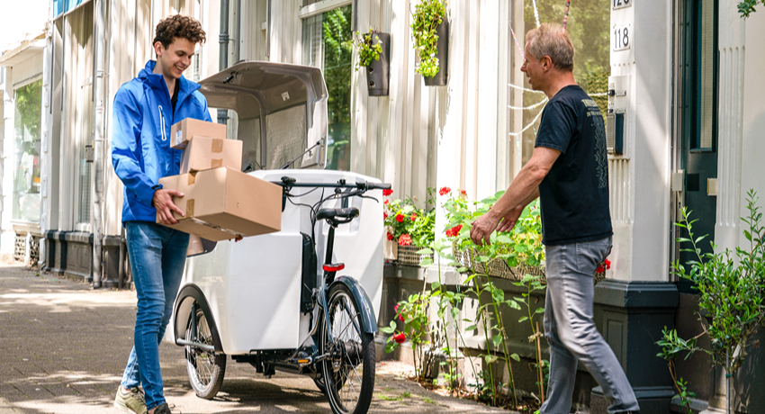 Un homme en vélo triporteur livre des colis à un habitant d'une ruelle fleurie.