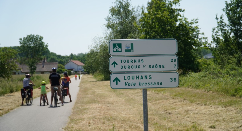 Un panneau indicateur sur la Voie Bressane avec au second plan une famille de randonneurs à vélo. 