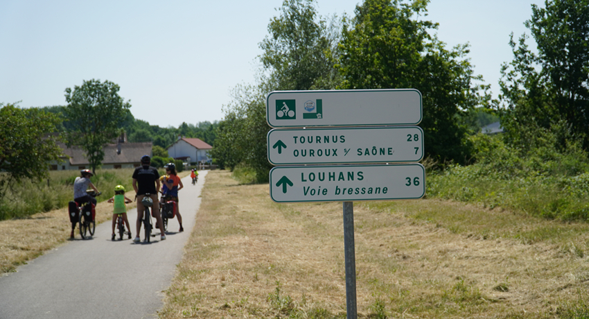 Un panneau indicateur sur la Voie Bressane avec au second plan une famille de randonneurs à vélo.