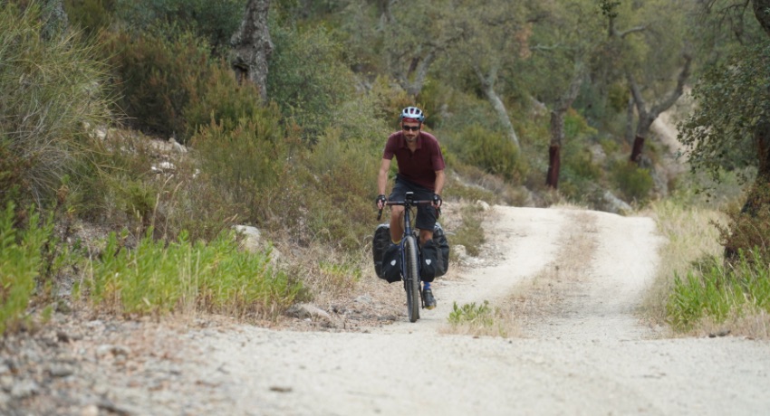 Un cycliste en gravel grimpe sur un chemin blanc qui traverse une forêt de chênes lièges.