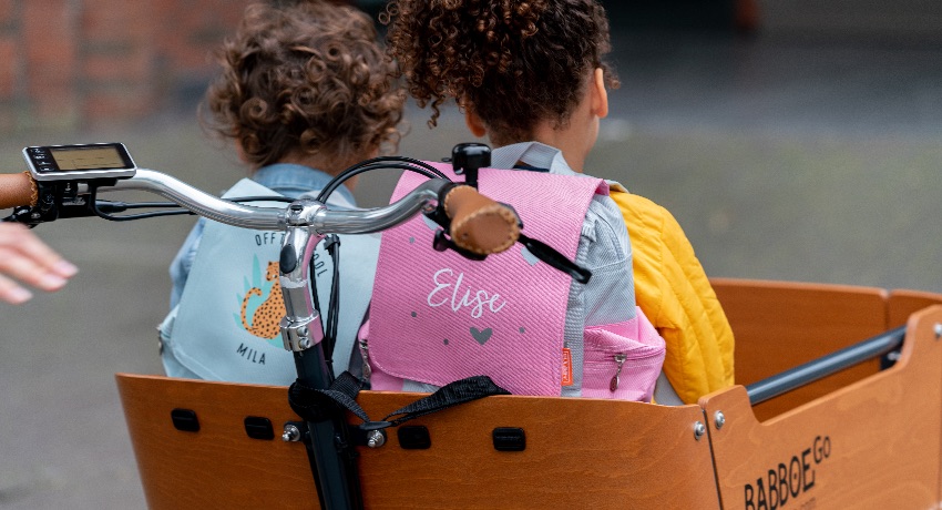 Deux petites filles de dos avec des sacs d'école installées dans la caisse d'un triporteur Babboe. 