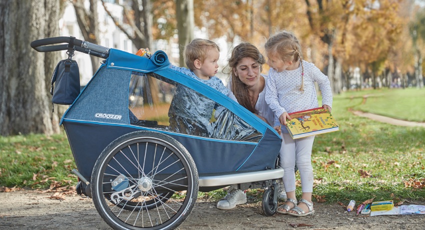 Un mère et ses deux jeunes enfants portant du matériel scolaire à côté d'une remorque vélo Croozer. 