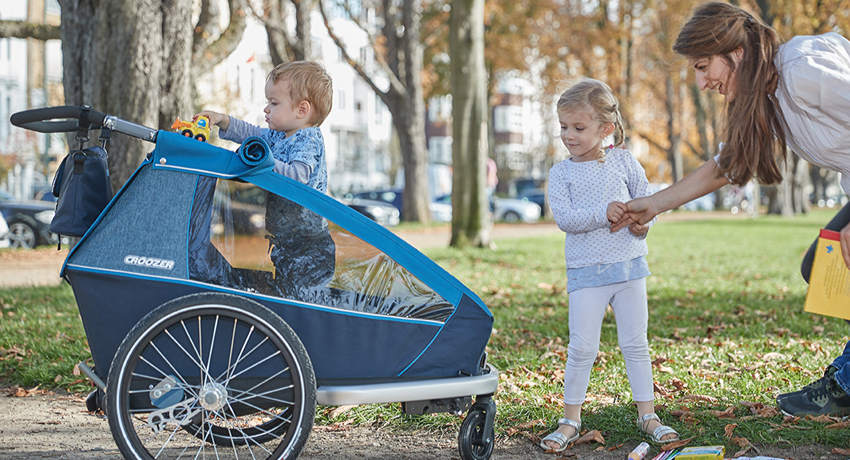 Une mère et ses deux jeunes enfants portant du matériel scolaire à côté d'une remorque vélo Croozer.