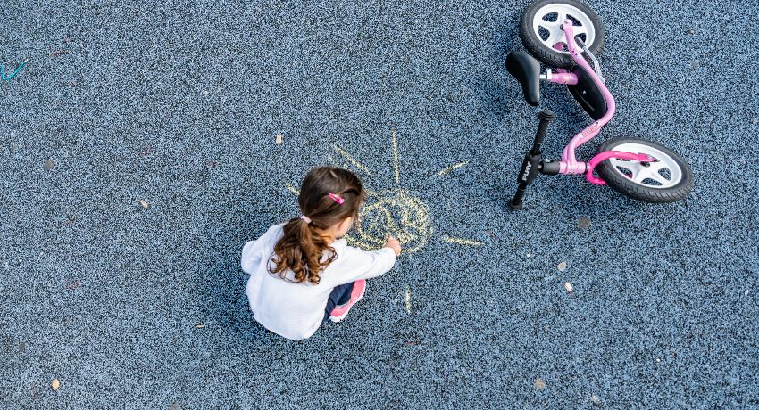 Une petite fille dessine un soleil à la craie dans la cour de récréation à côté de sa draisienne Puky.