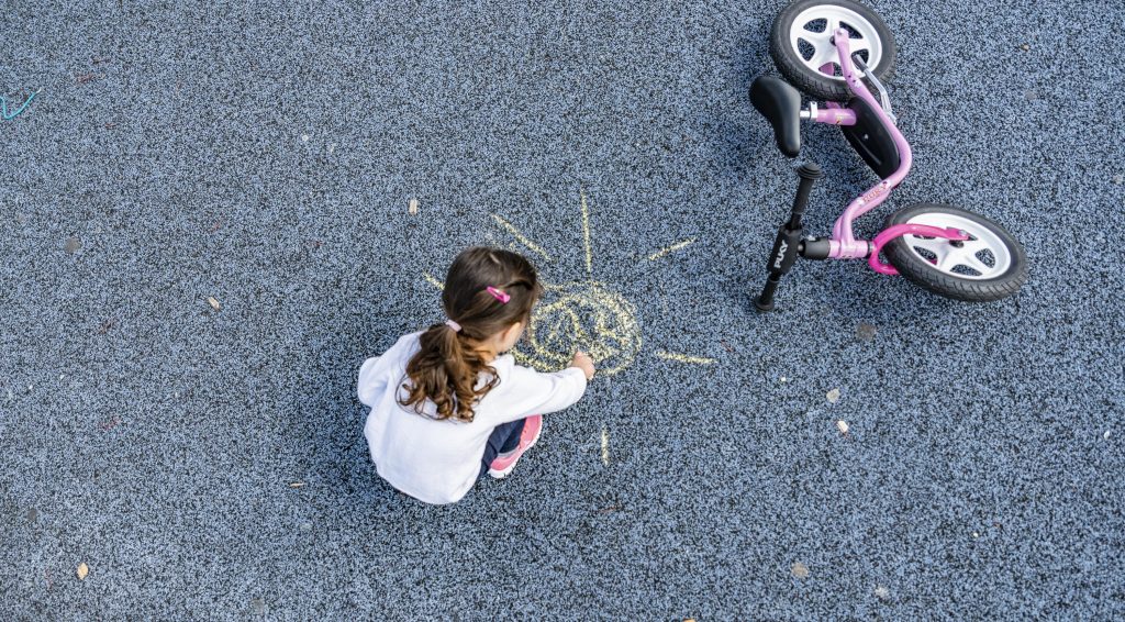 Une petite fille dessine un soleil à la craie dans la cour de récréation à côté de sa draisienne Puky.