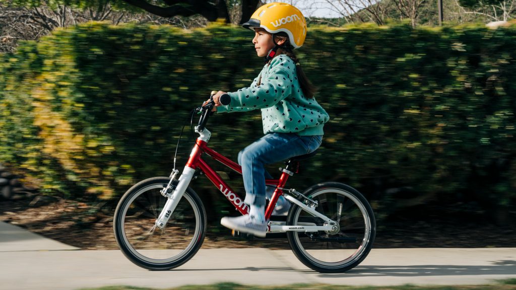 Une petite fille avec un casque jaune file sur un vélo woom Original 16 pouces rouge. 