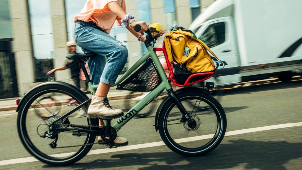 Une ado au guidon d'un vélo cargo pour enfant woom NOW transportant sur le porte-bagages avant un gros sac d'école jaune et d'autres accessoires scolaires. 