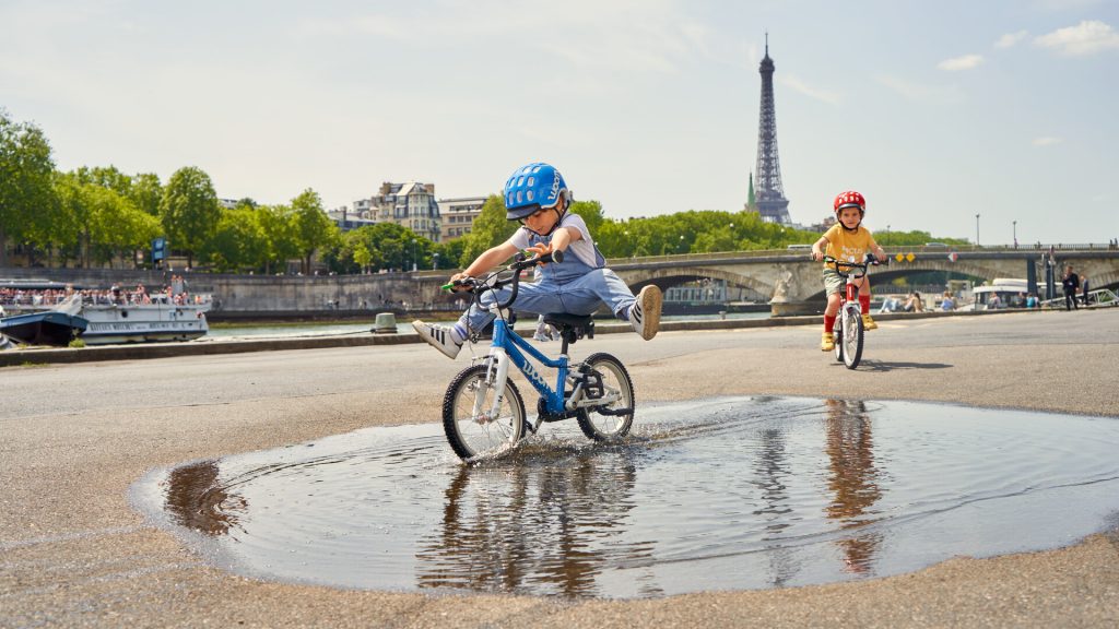 En bord de Seine avec la tour Eiffel en arrière plan, un enfant roule dans une flaque d'eau les jambes écartées au guidon de son vélo woom.