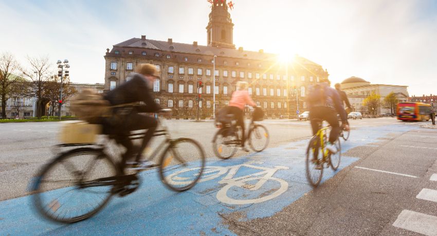 Des cyclistes progressent au coucher du soleil sur une voie cyclable urbaine matérialisée au sol par de la peinture bleue.