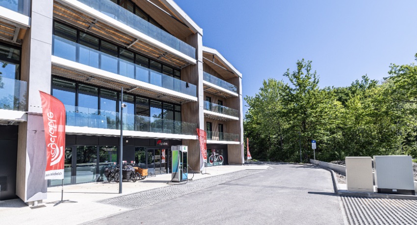Le bâtiment flambant neuf dans lequel est implanté le nouveau magasin de vélos Cyclable Grenoble-Meylan avec plusieurs place de parking et une borne de recharge pour véhicules électriques.