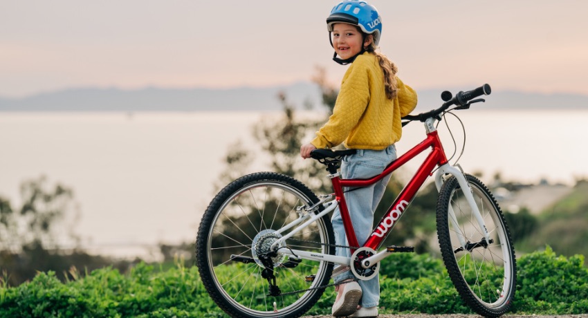 Une jeune fille avec un caque bleue observe la mer appuyée sur son vélo woom Original équipé de vitesses.  