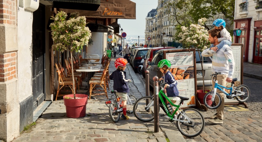 Un père de famille aide ses 3 enfants à traverser devant une boulangerie parisienne avec leurs vélos woom Original.