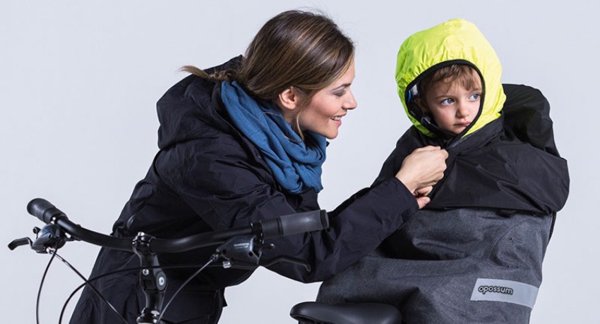 Une mère équipe son enfant installé dans un porte-bébé vélo de la cape de pluie enfant Tucano Urbano Opossum.