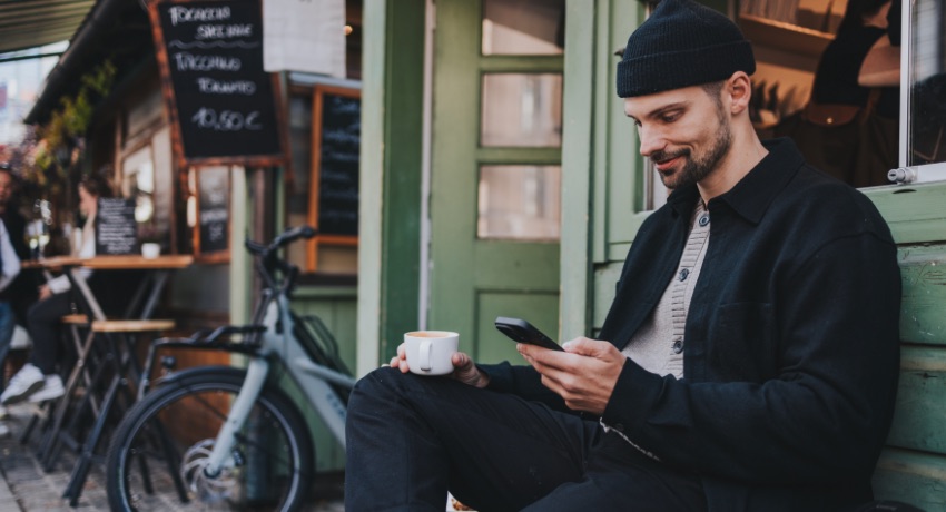 Un homme assis sur un banc avec un café devant son vélo cargo paramètre son application Bosch eBike Flow sur son téléphone.