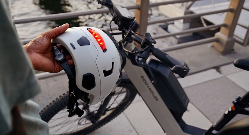 Une main tendue tient un caque Bern muni d'un dispositif d'éclairage intégré devant un VAE Riese & Müller.