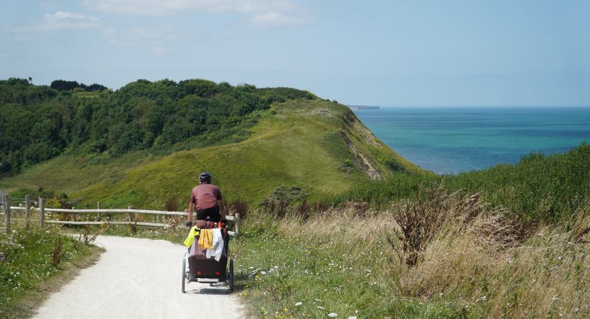 Un cycliste avec une remorque descend une voie cyclable en gravier dominant la mer turquoise et des falaises verdoyantes en Normandie.