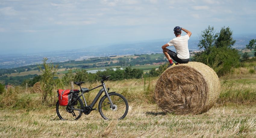 Un cycliste assis sur une botte de foin contemple l'horizon en contrebas. A côté, attend un VAE avec des sacoches de voyage.