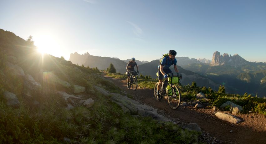 Deux cyclistes en configuration bikepacking progressent sur un sentier de montagne au soleil couchant.