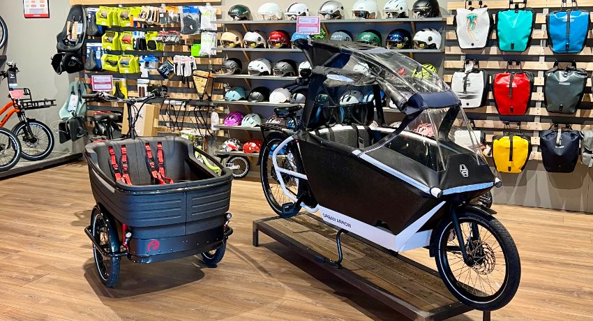 Un vélo cargo longtail et un triporteur dans le showroom du magasin de vélo Cyclable Lausanne Malley. Au second plan, un portant avec des sacoches colorées et des casques.