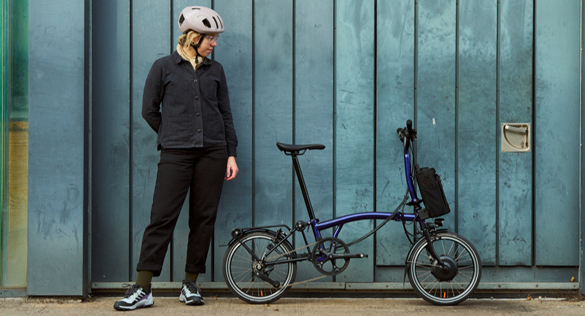 Une femme pose devant un mur de planches bleues aux côtés d'un Brompton électrique P Line.