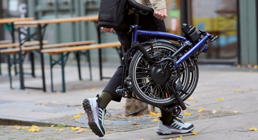 Une personne marche en ville avec à la main un Brompton électrique plié et à l'épaule la sacoche contenant la batterie du vélo.
