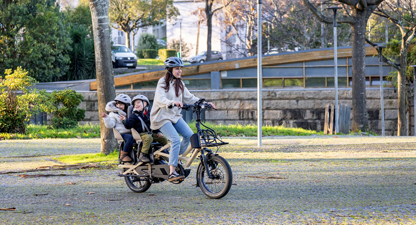 Une femme transporte deux enfants en bas âge sur un longtail Tern GSD Gen3 à travers un parc urbain boisé.