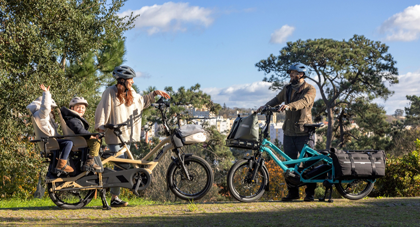 Dans un parc urbain arboré deux Tern GSD Gen 3 se font face. L'un avec une femme et deux enfants sur le porte bagages. L'autre avec de volumineuses sacoches arrière tenu par un homme.
